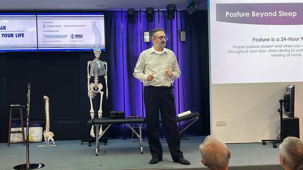 Y. Zafer delivering a posture education talk at an event co-hosted by Chiropractic Specialty Center® and IMU, with a skeleton model and posture slides in the background.